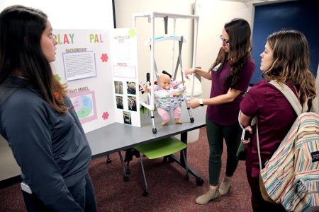 OTA student Tamiya Vanhook-Davis  (right) shows fellow student Hannah Cook (left) how to cook using an adaptive tray sOTA students Laurel Pellant and Jordan Epperly (left to right) demonstrate a device for first-year OTA student Hali Creasey (right) that will help the parents of an infant born with Spastic Quadriplegia complete everyday tasks.he created.