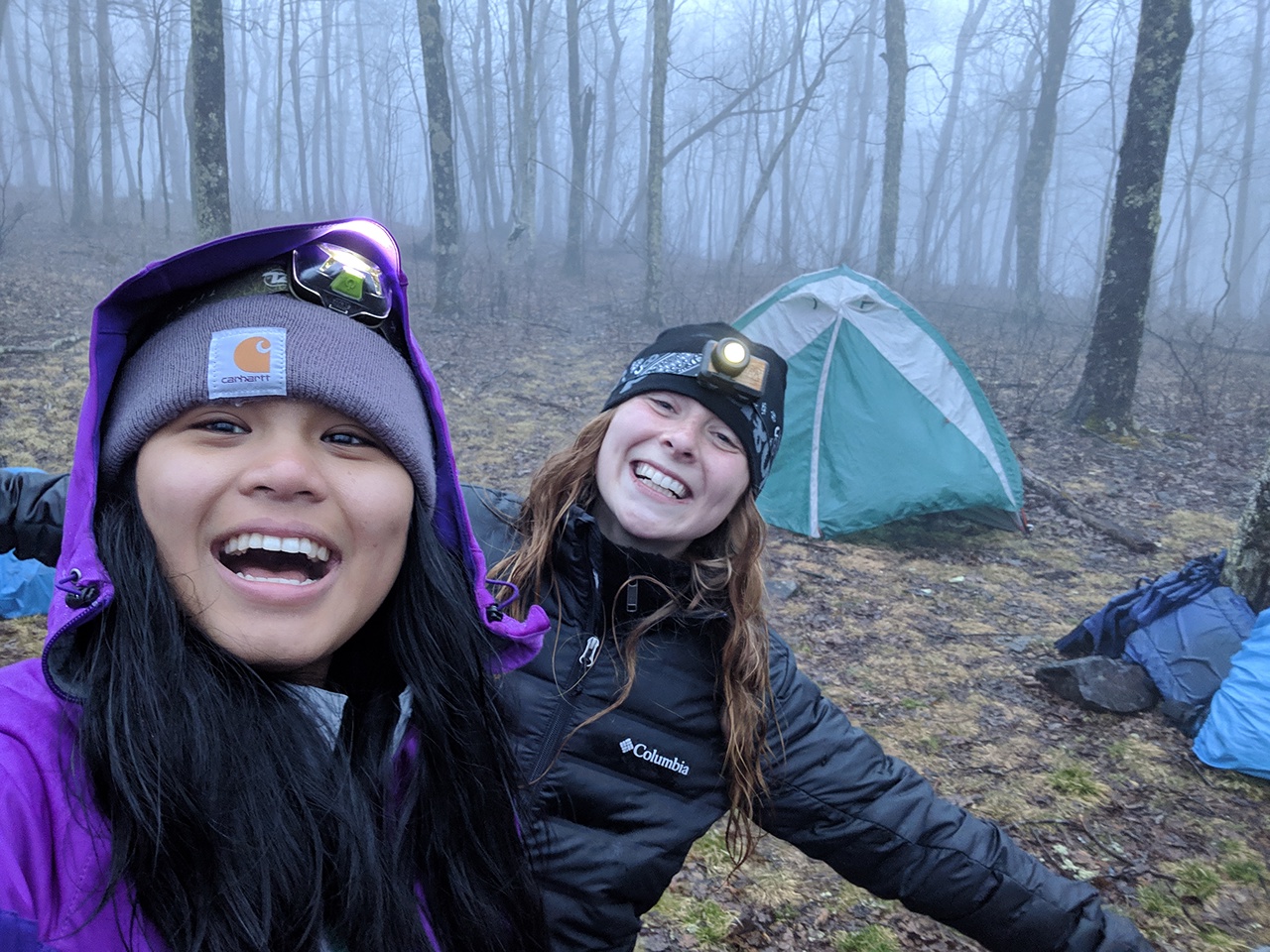 Two female students smiling in front a a tent in the outdoors.