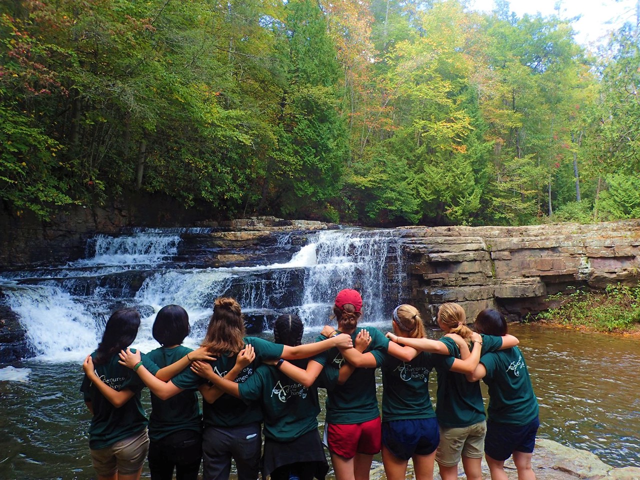 A group of 8 students stand together, arms linked, in front of a small waterfall.