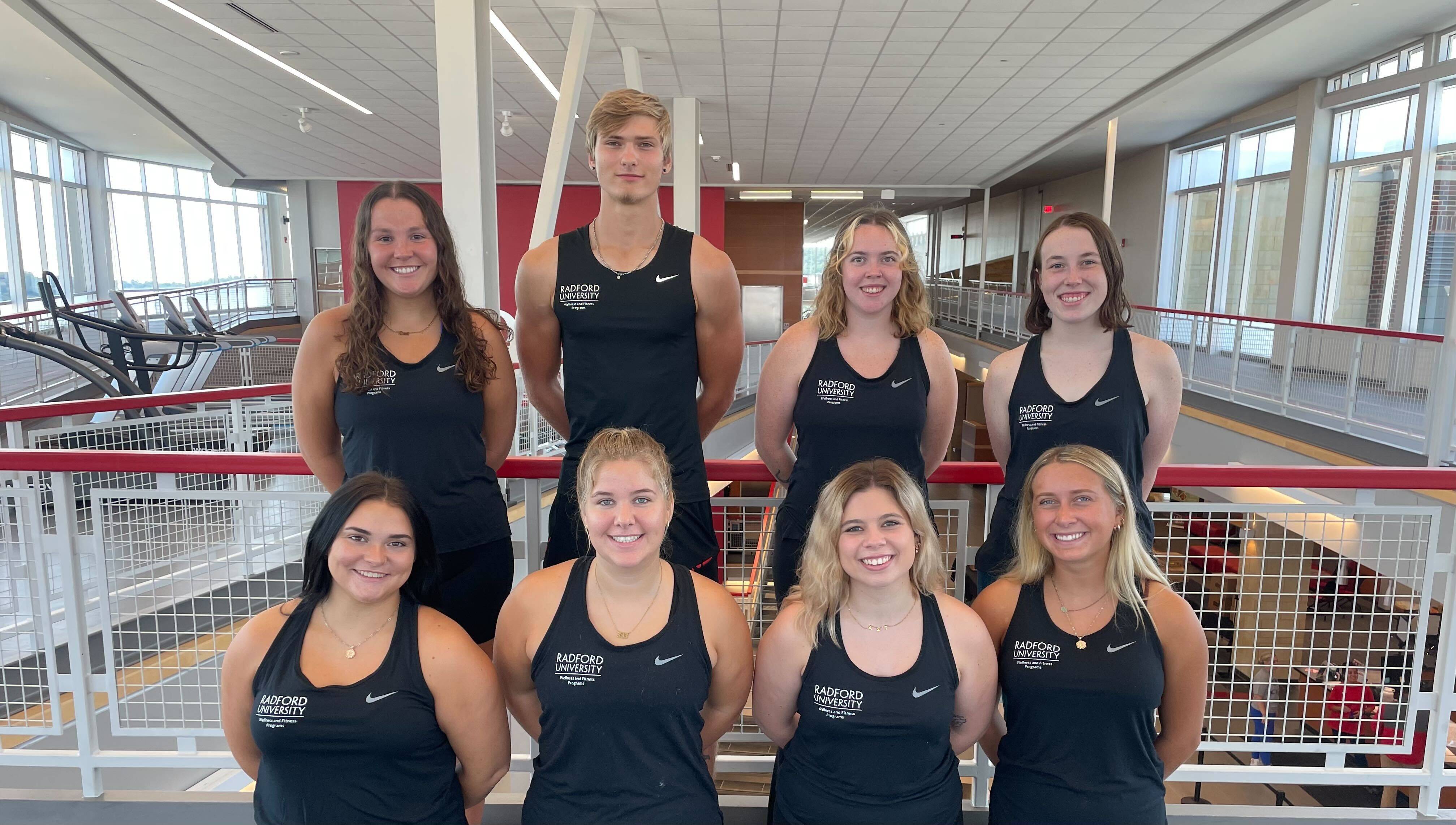 A group of eight individuals wearing matching black athletic tank tops stands inside a spacious indoor facility. They are positioned on two levels of a tiered arrangement, with five individuals in the back row and three in the front row. The setting features large windows, natural light, and red-and-white railings overlooking exercise equipment and an open fitness area. The tank tops display a logo on the left side that reads “Radford University Student Recreation & Wellness.”