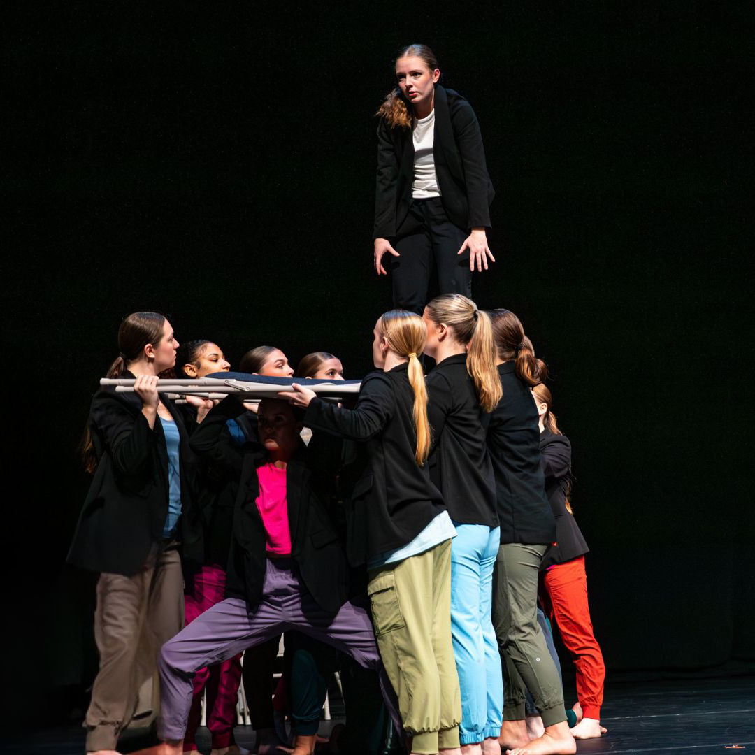 Dancers in black blazers lift a dancer using folding chairs