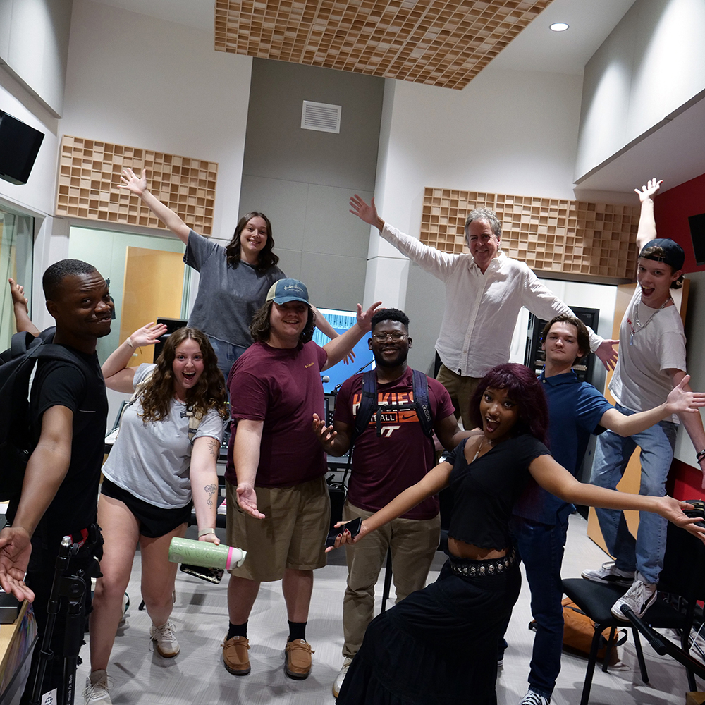 Director David Rivers poses with students from Radford University's Commercial Music Ensemble inside the recording studio at the Artis Center. The group stands together in the studio with arms outstretched, smiling toward the camera during a recording session.