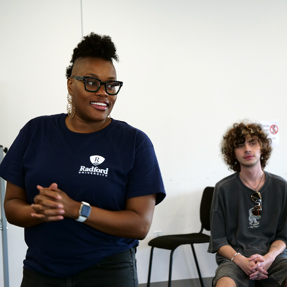 Woman in glasses wearing a Radford University T-shirt standing in a room, with a curly-haired young man seated behind her.